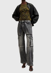 Person som bär en svart skinnjacka, grönt leopardmönstrat linne, grå cargojins och svarta sandaler med öppen tå och hög klack, stående med händerna i fickorna.