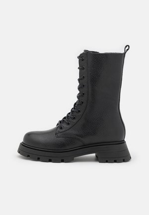 Botte de combat en cuir noir avec finition texturée, devant à lacets, semelle épaisse à crampons et languette de tirage à l'arrière.