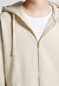 Felpa beige con zip realizzata in tessuto morbido, dotata di cappuccio con cordino, tasche frontali e una texture liscia. Indossata sopra una maglietta bianca aderente.