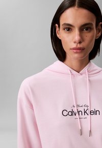 Γυναίκα με κοντά σκούρα μαλλιά που φοράει ροζ ανοιχτό φούτερ με κουκούλα, με την επιγραφή "New York City Calvin Klein" στο μπροστινό μέρος, με ουδέτερη έκφραση, γκρι φόντο.