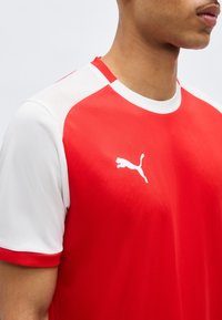 Κόκκινο και λευκό αθλητικό πουκάμισο με κοντά μανίκια και το λογότυπο της Puma στο στήθος, φορεμένο από ένα άτομο που φαίνεται από τον λαιμό μέχρι το πάνω μέρος του βραχίονα, με φόντο απλό.