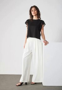 Crop top noir avec des manches flottantes transparentes, associé à un pantalon large blanc et des sandales à lanières noires. Design simple et élégant.