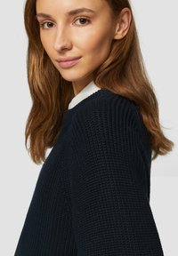 Maglione lavorato a maglia di colore blu navy con un motivo testurizzato; presenta un colletto bianco che spunta da sotto; modella con lunghi capelli castani e mossi.