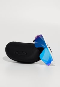 Occhiali da sole sportivi con montatura bianca e lenti blu sfumate, appoggiati su un astuccio rigido nero con un logo circolare.