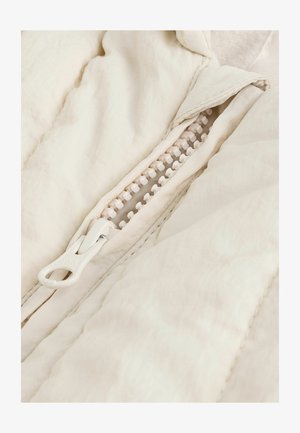 Kvadri muretähega kreemjas jope, millel on plastist tõmblukk koos valge tõmbluku tõmbeosaga. Sile kangatekstuur ja peened õmblusdetailid on nähtavad.