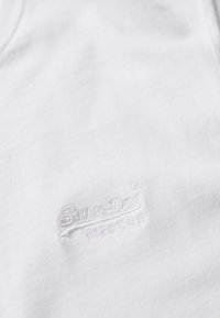 Camiseta de algodón blanca con escote redondo y un logo texturizado y en relieve en color gris claro, con el nombre de la marca "SuperDry" prominentemente exhibido.