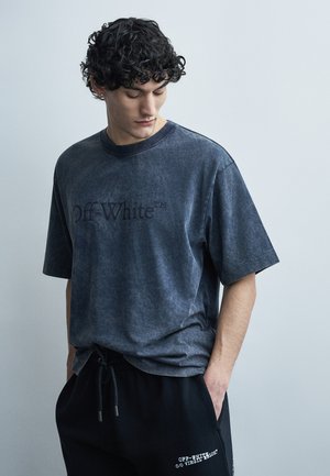 Mladý muž s kučeravými vlasmi nosiaci vyblednuté tmavosivé tričko Off-White a čierne tepláky Off-White, s rukami v kapse.