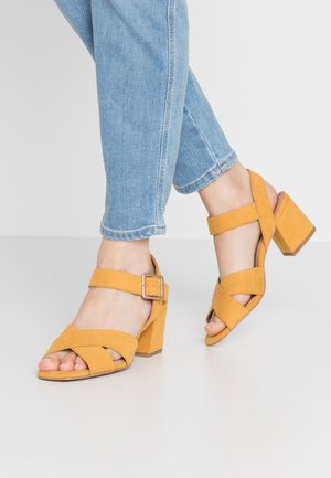 Beine in hellblauen Jeans, die senfgelbe Sandalen mit Blockabsatz, Knöchelriemen und Schnallen auf einem einfarbigen hellen Hintergrund tragen.