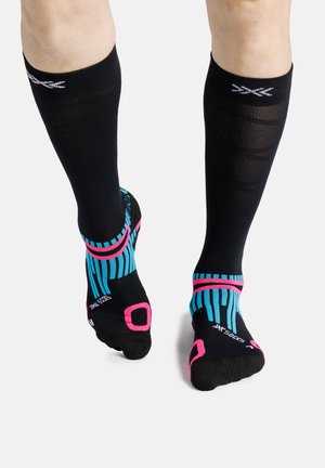 Benenen met zwarte kniehoge compressiekousen met blauwe en roze patronen op de voeten, staand op een witte achtergrond.