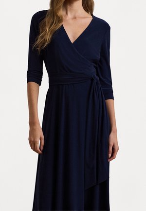 Vrouw draagt een marineblauwe, lange mouwen wikkeljurk met een V-hals en een gestrikte tailleband, getoond van hals tot halverwege de dij.