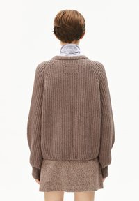 Person von hinten gezeigt, trägt einen grob gestrickten taupefarbenen Pullover über einem braunen Tweed-Rock und ein lavendelfarbenes Rollkragenshirt.