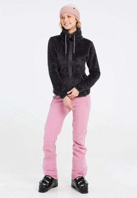 Zwarte fleece jack met hoge kraag, roze skibroek en zwarte skischoenen. Lichtroze gebreide beanie. Glad materiaal en een relaxte pasvorm.