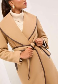 Wollmischung-Mantel aus Kamelhaar mit braunen Lederdetails, großem Kragen und tailliertem Gürtel; kombiniert mit einem leichten Pullover und auffälligen Ohrringen.