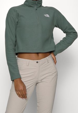 Žena v zeleném krátkém pulovru se čtvrtinovým zipem a béžových kalhotách, jedna ruka na kyčli, druhá volně podél těla.