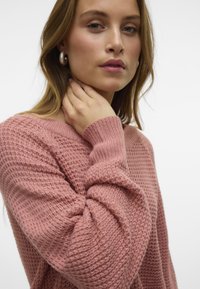 Pull en tricot rose avec un motif texturé, un col large et des poignets côtelés. Le modèle se tient le cou, mettant en valeur de belles boucles d'oreilles pendantes.