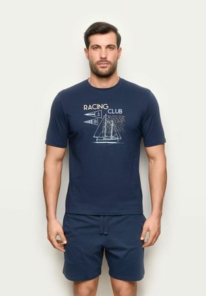Mann trägt dunkelblaues T-Shirt mit Segelboot und Schriftzug „Racing Club“ kombiniert mit passenden dunkelblauen Shorts, steht vor einfarbigem Hintergrund.
