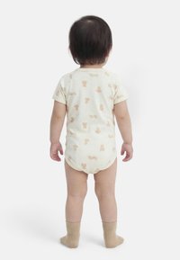 Νήπιο που στέκεται γυρισμένο προς τα πίσω, φοράει κοντομάνικο εκρού φορμάκι με στάμπες αρκουδάκι και μπεζ κάλτσες σε λευκό φόντο.