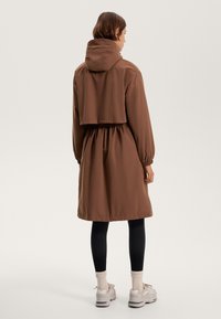 Bruine, lichte, met capuchon uitgeruste jas met een getailleerde taille, raglanmouwen en een cropped bovenstuk. Draagt over zwarte leggings en sneakers.