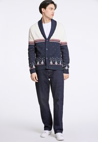 Mörkblå cardigan med krämfärgad sjalkrage, mönstrade snöflingor och träd. Bärs med mörka jeans och vita skor, har knapprad fram.