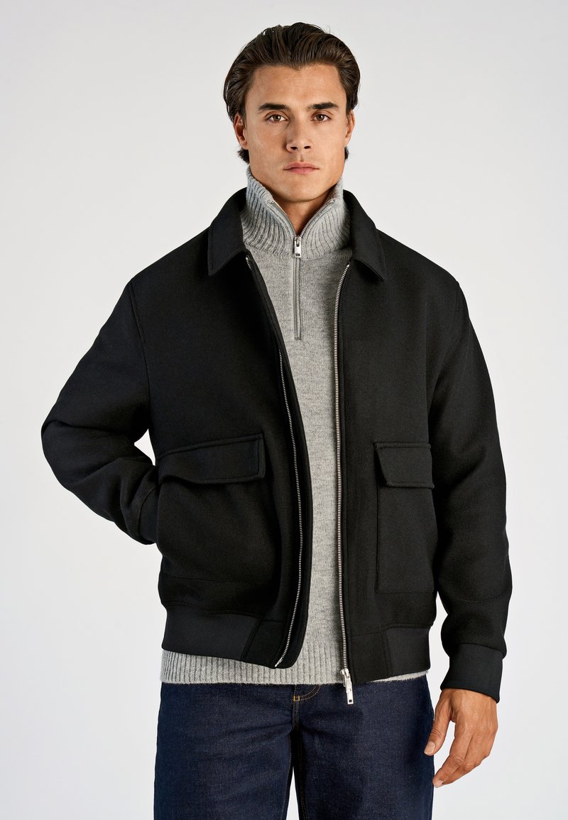 Svart bomberjacka med dragkedja, två stora framfickor och ribbad nederkant. Buren över en ljusgrå polotröja.