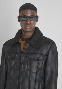 Jonge man in een zwart schapenvachten jasje over een bruine trui, met rechthoekige zwarte zonnebril, staand tegen een neutrale achtergrond.