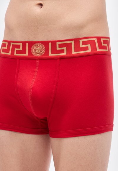 Κόκκινα βαμβακερά ανδρικά μποξεράκια με χρυσή ζώνη μέσης διακοσμημένη με σχέδιο ελληνικού κλειδιού και λογότυπο με μετάλλιο. Λείο ύφασμα, εφαρμοστή γραμμή.