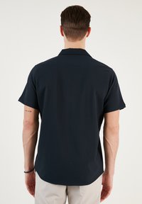 Tengerészeti, rövid ujjú ing lapos gallérral, sima textúrával és laza szabással, oldalsó hasítékkal az alján. Csak hátsó nézet.
