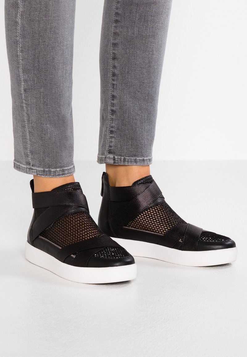 Pieds chaussés de baskets montantes noires avec des panneaux en mesh et de larges sangles, associées à un jean gris coupé au-dessus de la cheville.