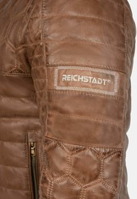 Bruine leren jas met een getextureerd quiltpatroon en hexagonale vormen. Voorzien van een ritssluiting en een label met de borduurlijke details van "REICHSTADT".