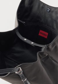 Μαύρη δερμάτινη τσάντα ώμου με ρυθμιζόμενο λουρί, ανοιχτό πάνω μέρος που δείχνει εσωτερική τσέπη με φερμουάρ και κόκκινη ετικέτα "HUGO".