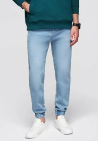 Jogger jeans de color azul claro con puños elásticos, detalles desgastados y un corte relajado, combinados con zapatillas blancas y un sudadera verde.