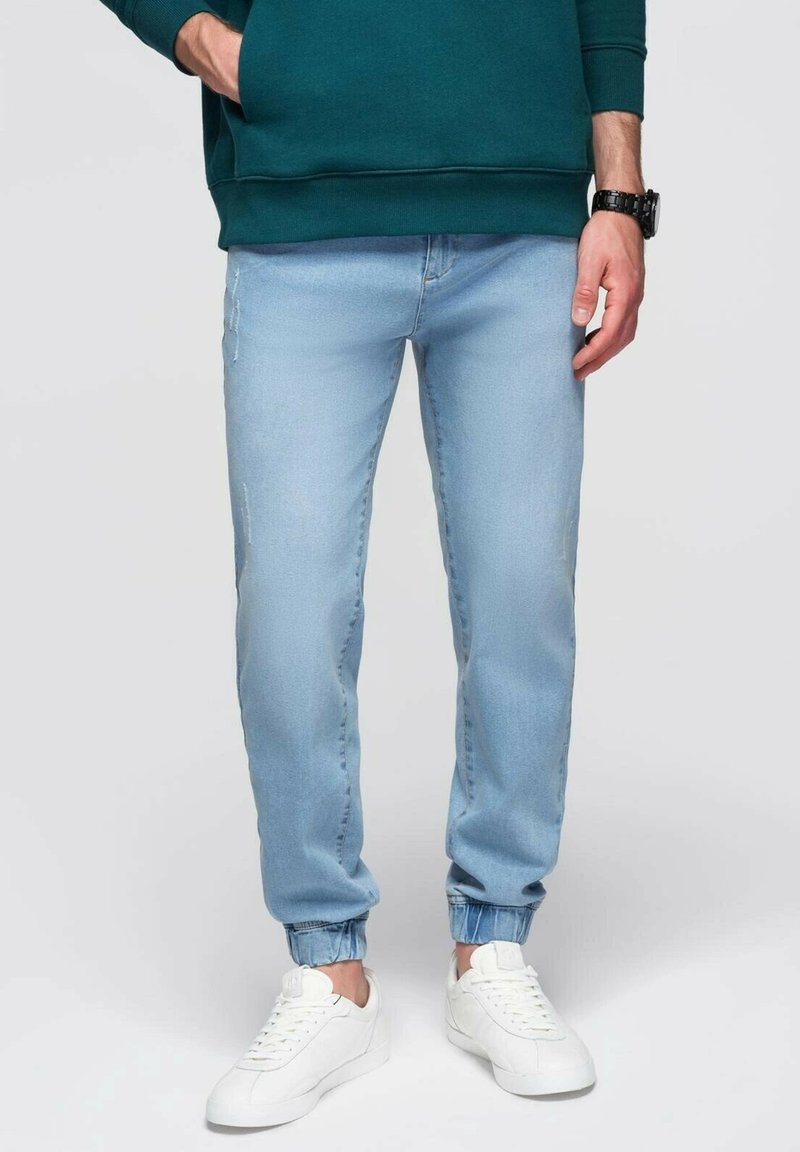 Jogger jeans de color azul claro con puños elásticos, detalles desgastados y un corte relajado, combinados con zapatillas blancas y un sudadera verde.