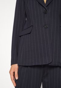 Veste de costume bleu marine à fines rayures avec deux boutons, poches à rabat et pantalon assorti à fines rayures sur le torse et la main d'une personne.