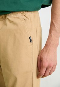 Beige stofbukser med elastisk talje, der har en sidelomme og et lille sort mærke. Glat tekstur med subtile syningsdetaljer.