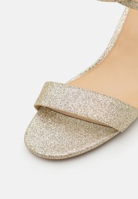 Sandale cu glitter auriu, având o bandă largă pe picior și un design cu vârful descoperit. Talpă netedă, plată, cu un luciu metalic.
