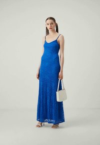 Vestido maxi azul rendado com alças finas, design ajustado, intricado padrão de renda e uma bolsa branca. Modelo a usar sandálias.