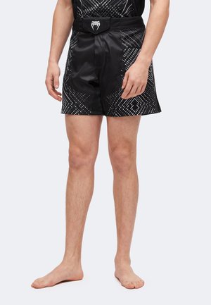 Hombre con pantalones cortos deportivos negros con patrones geométricos blancos y una cintura con un logo de serpiente, de pie descalzo.