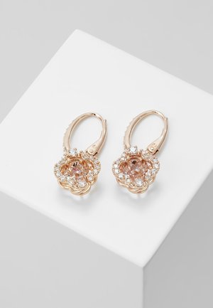 Boucles d'oreilles créoles en or rose avec des montures en forme de fleurs, ornées de pierres roses centrales entourées de petits cristaux clairs, posées sur une surface blanche.