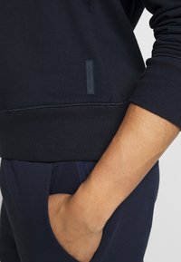 Person trägt einen marineblauen Pullover und eine Hose, die Hand ruht in der Hosentasche, sichtbares Michael Kors Logo auf dem Pullover.