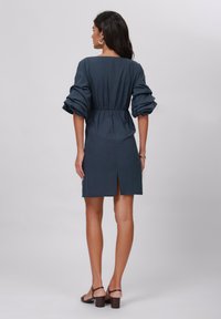 Robe bleu foncé avec un décolleté carré, des manches bouffantes, une taille élastique et une fente au dos. Tissu lisse avec une silhouette ajustée.