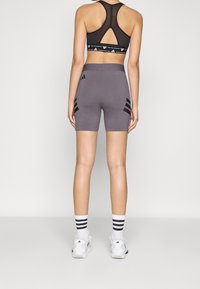 Žena oblečená v černé síťované sportovní podprsence, šedých cyklistických šortkách Adidas, bílých ponožkách s černými pruhy a bílých teniskách Adidas, stojí s obličejem k divákovi.