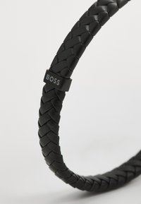 Pulsera de cuero trenzado negra con un pequeño acento de metal negro que lleva la palabra "BOSS" en plata, mostrando una superficie lisa y texturizada.