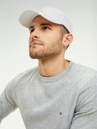 Skægget mand iført en hvid kasket og en lysegrå sweatshirt, der ser eftertænksomt opad mod en ensfarvet lys baggrund.