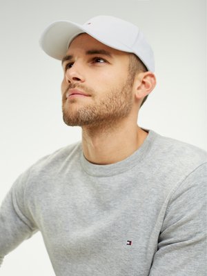 Skægget mand iført en hvid kasket og en lysegrå sweatshirt, der ser eftertænksomt opad mod en ensfarvet lys baggrund.