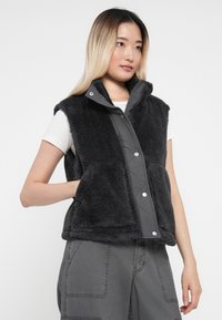 Melnā veste, kas izgatavota no mīksta auduma, ar gludu quilted priekšpusi, pogām, kas aizver vestes priekšpusi, un divām priekšējām kabatām; apvienota ar pelēkiem biksēm.