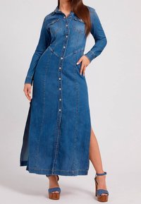 Kvinna i lång blå denimklänning med knappar, långa ärmar och sidssprund, i kombination med blå klackskor.