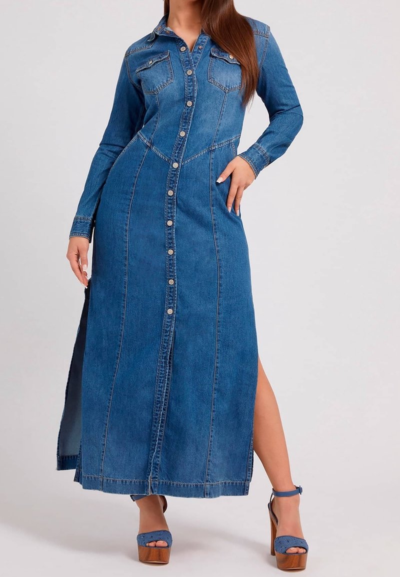 Kvinna i lång blå denimklänning med knappar, långa ärmar och sidssprund, i kombination med blå klackskor.