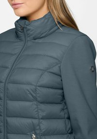 Frau, die eine blaue gesteppte Jacke mit Reißverschluss und Stoffärmeln trägt, von den Schultern bis zur Mitte des Oberkörpers sichtbar, nach rechts gewandt.