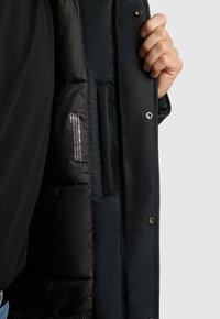 Czarna, pikowana kurtka z błyszczącym wnętrzem, zatrzaskami i widoczną metką wewnątrz. Dłoń chwytająca krawędź kurtki w pobliżu kieszeni.