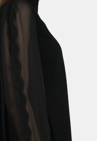 Top nero con maniche nere trasparenti, caratterizzato da un dettaglio in pizzo a scallop lungo la cucitura, che crea un effetto di texture a strati.